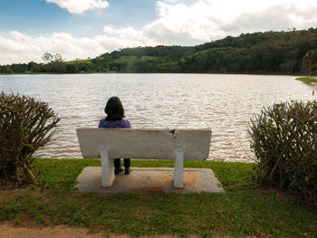 Rear view of man sitting on bench by lake against sky