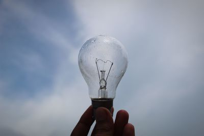 Close-up of hand holding light bulb against sky