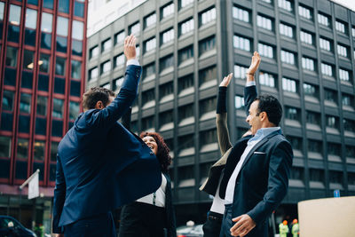 Cheerful multi-ethnic business colleagues standing with raised hands against building in city