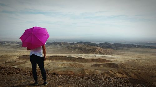 Rear view of person with umbrella standing on cliff against cloudy sky