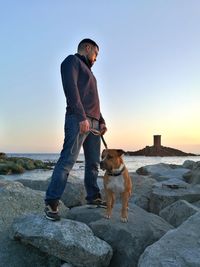 Man with dog standing on rock against sky during sunset