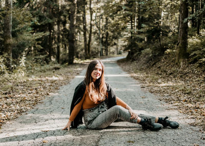 Beautiful young woman in autumn clothes sitting in middle of empty road in forest.