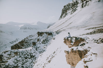 Unrecognizable cosmonaut wearing white spacesuit sitting on edge of rocky mountain in winter and admiring amazing landscape in svalbard