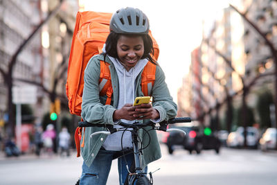 Side view of positive african american female cyclist with thermo backpack standing on paved sidewalk while checking order information on cellphone