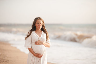 Smiling pregnant woman wear white dress hol tummy walk at beach over sea nature background outdoors.