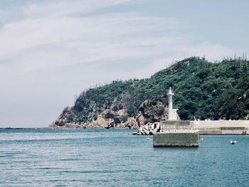 The pier in the port with a small lighthouse against the blue ocean, sky, and forest.