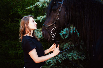 Side view of young woman standing with horse by trees
