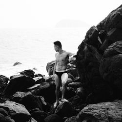 Rear view of shirtless man standing on rock at beach