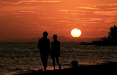 Silhouette people standing at beach against orange sky