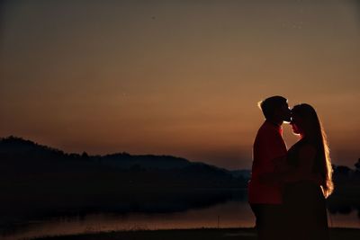 Side view of young couple kissing at lakeshore against sky during sunset