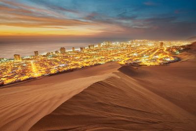 Desert by illuminated cityscape and sea against sky during sunset