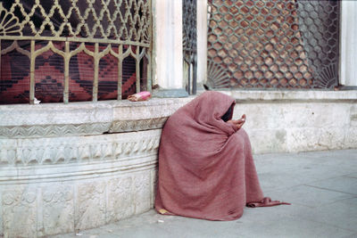 Full length of man covering in blanket sitting on footpath