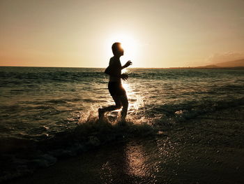 Boy running on shore at beach against sky during sunset