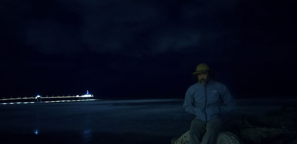 Man sitting in illuminated sea against sky at night