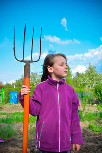 Girl looking away while holding gardening fork against blue sky in yard