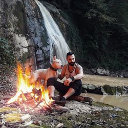 Man sitting by bonfire against waterfall