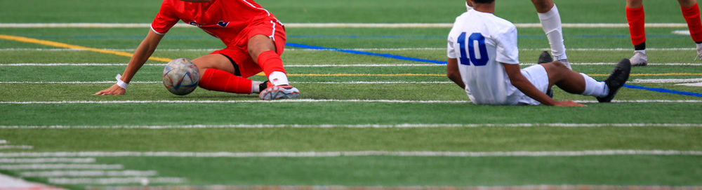 Soccer players falling while chasing a soccer ball during a game on a turf field.