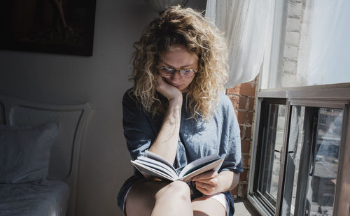 Woman reading book while sitting on window sill at home
