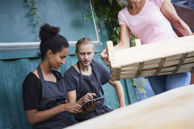 Male and female gardener using tablet computer while senior woman carrying crate in yard