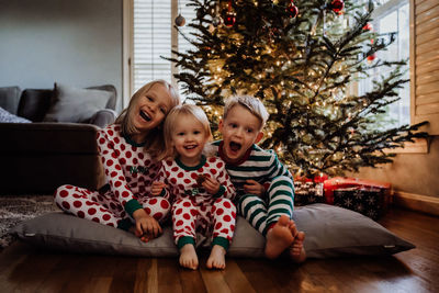 Siblings in costume sitting against christmas tree at home