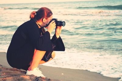 Full length side view of woman photographing while crouching on shore at beach during sunset