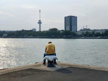 Rear view of man sitting on river in city against sky