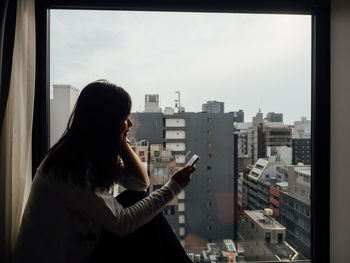 Woman using mobile phone while sitting by glass window