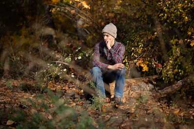 Sad teen boy in beanie, flannel, and jeans sits on log in woods.