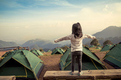 Rear view of girl with arms outstretched standing on retaining wall against sky at campsite