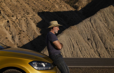 Adult man in cowboy hat standing against car on desert against mountain. almeria, spain