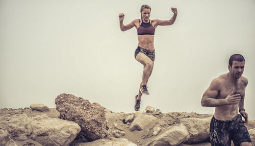 Man running while woman jumping at dirt road against clear sky