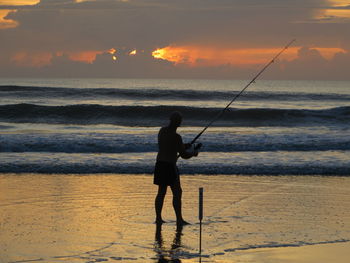 Man fishing in sea against sunset sky