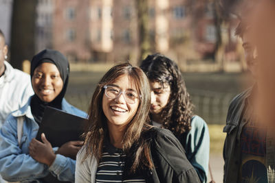 Portrait of smiling young female student with friends at university campus on sunny day