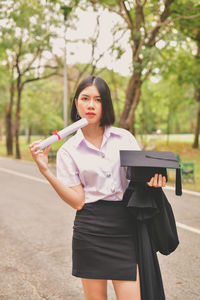 Portrait of young woman holding mortarboard while standing on road at park