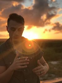 Young man holding cigarette standing against sun during sunset
