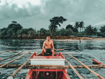 Portrait of shirtless man sitting on boat in river against sky