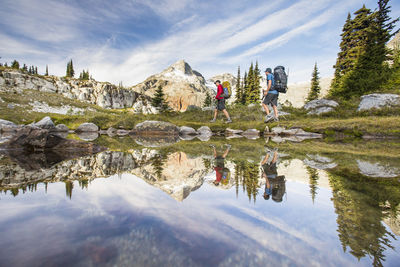 Reflection of backpackers hiking beside alpine lake in mountains.