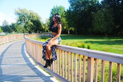 Full length portrait of woman sitting on railing against trees