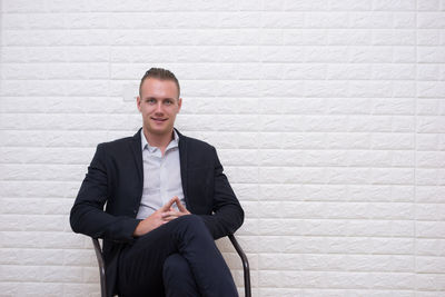 Portrait of businessman sitting on chair against white brick wall