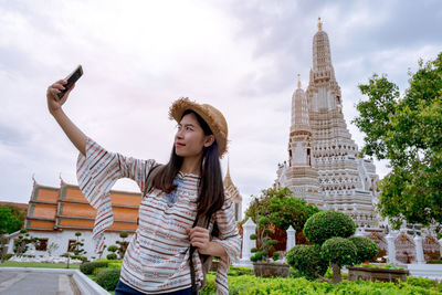 Young woman taking selfie while standing by temple against cloudy sky