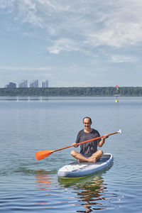 Asian older man sitting on sup board, rowing with oar on calm lake. good moments concept.