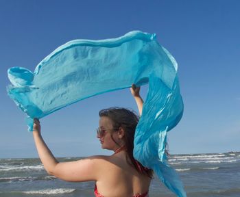Rear view of girl holding textile at beach against blue sky