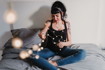 Pregnant young woman sitting with string lights on bed at home