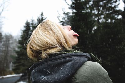 Side view of young woman in warm clothing sticking out tongue while standing against trees at park
