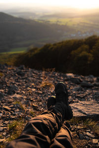 Low section of man sitting on rock against sky during sunset