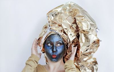 Close-up of young woman with face paint covering hair in crumpled golden foil against white background