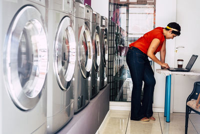 Side view of woman using laptop standing in laundromat