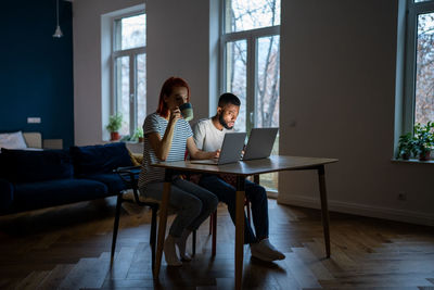 Young modern freelancing couple sitting at table in living room working from home together