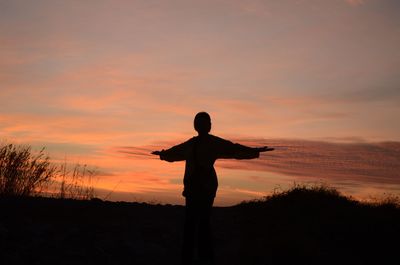 Silhouette woman with arms outstretched standing on landscape against sky during sunset