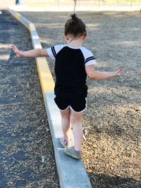 Rear view of girl balancing while walking on retaining wall by road